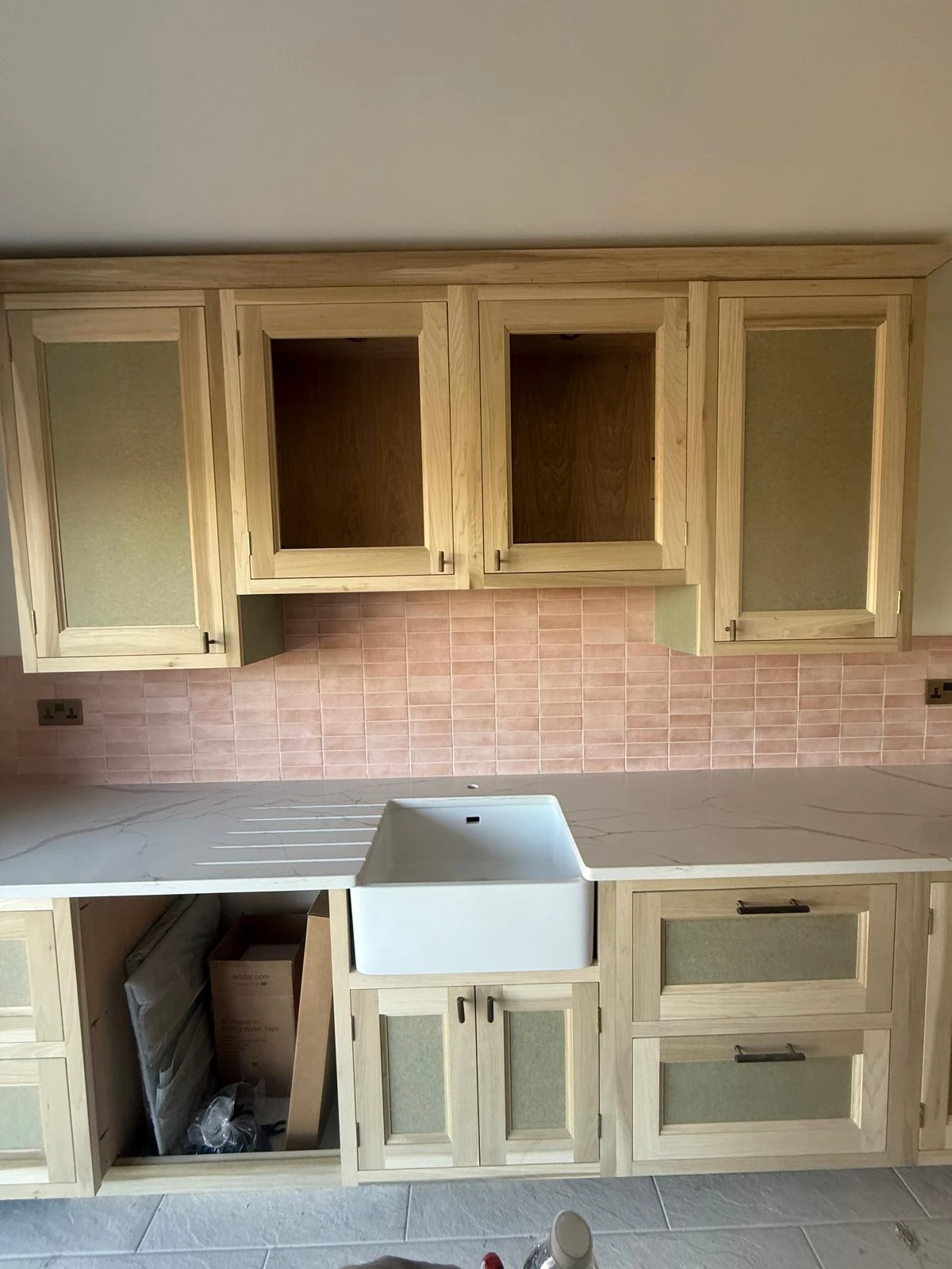 A partially finished kitchen featuring wooden cabinets, a white sink, and pink tiled backsplash. Some cabinets are open showing storage.