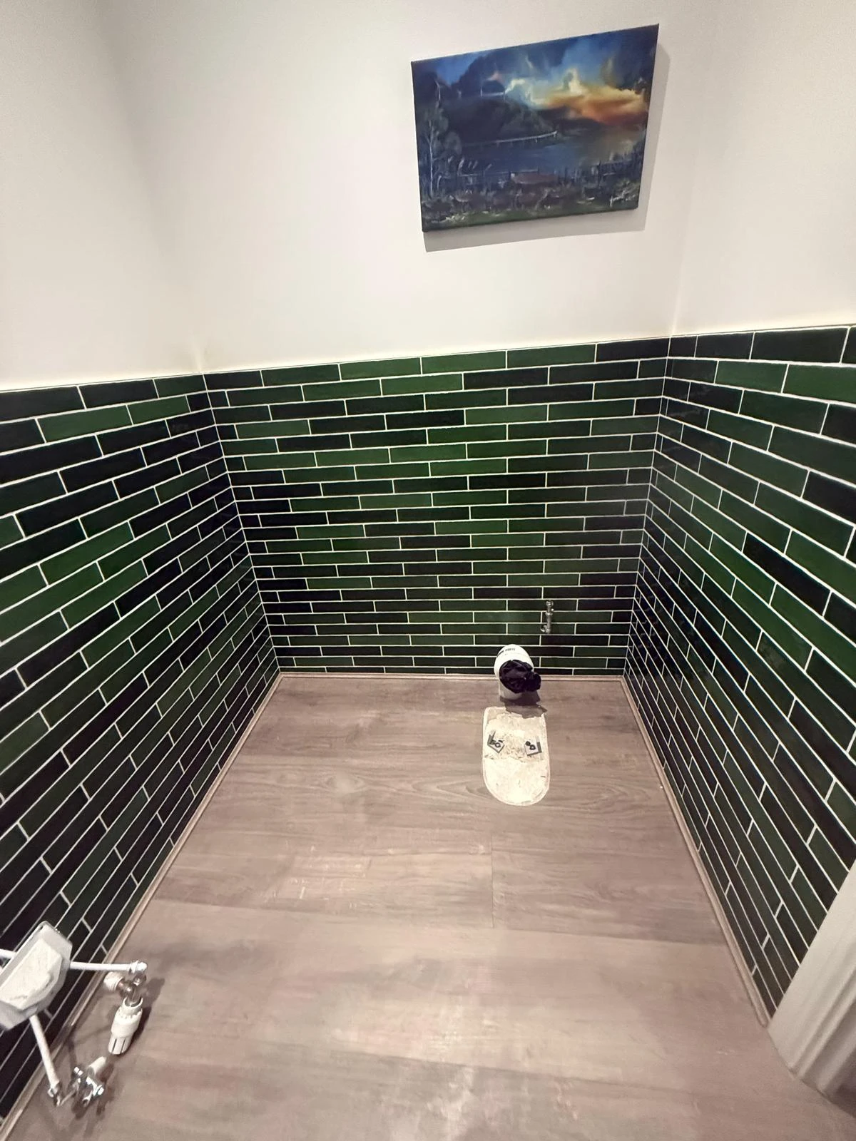 Small bathroom corner with dark green tiled walls, wooden floor, and a toilet setup. A landscape painting hangs above.