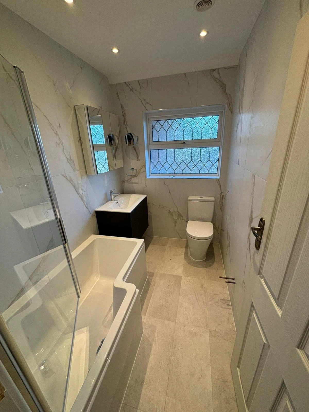 Modern bathroom with a bathtub and shower, sink with cabinet, toilet, and a window featuring decorative glass. Brightly lit with neutral tones.