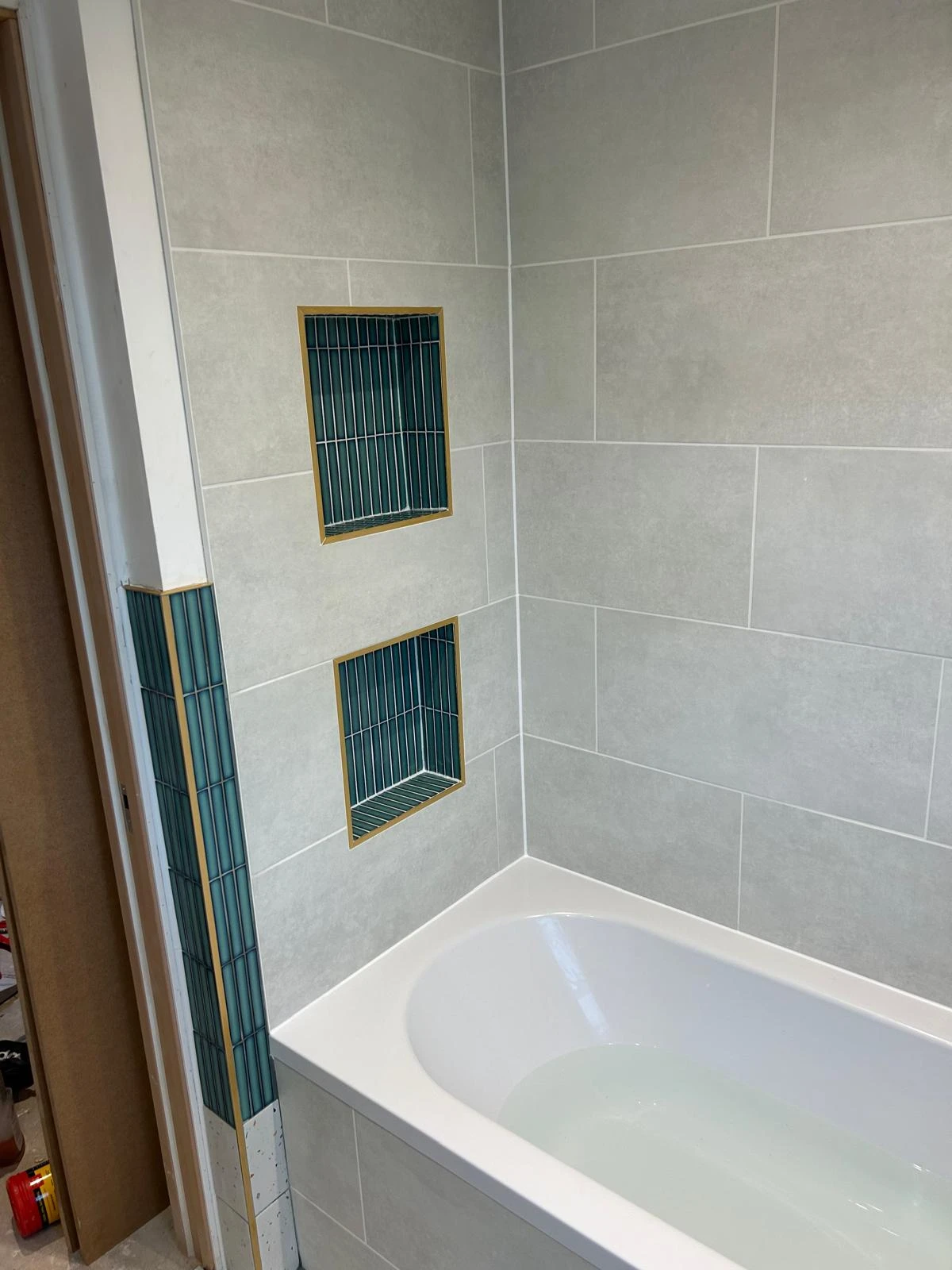 Modern bathroom featuring a white bathtub, textured grey tiles, and two wall niches with teal and gold accents.