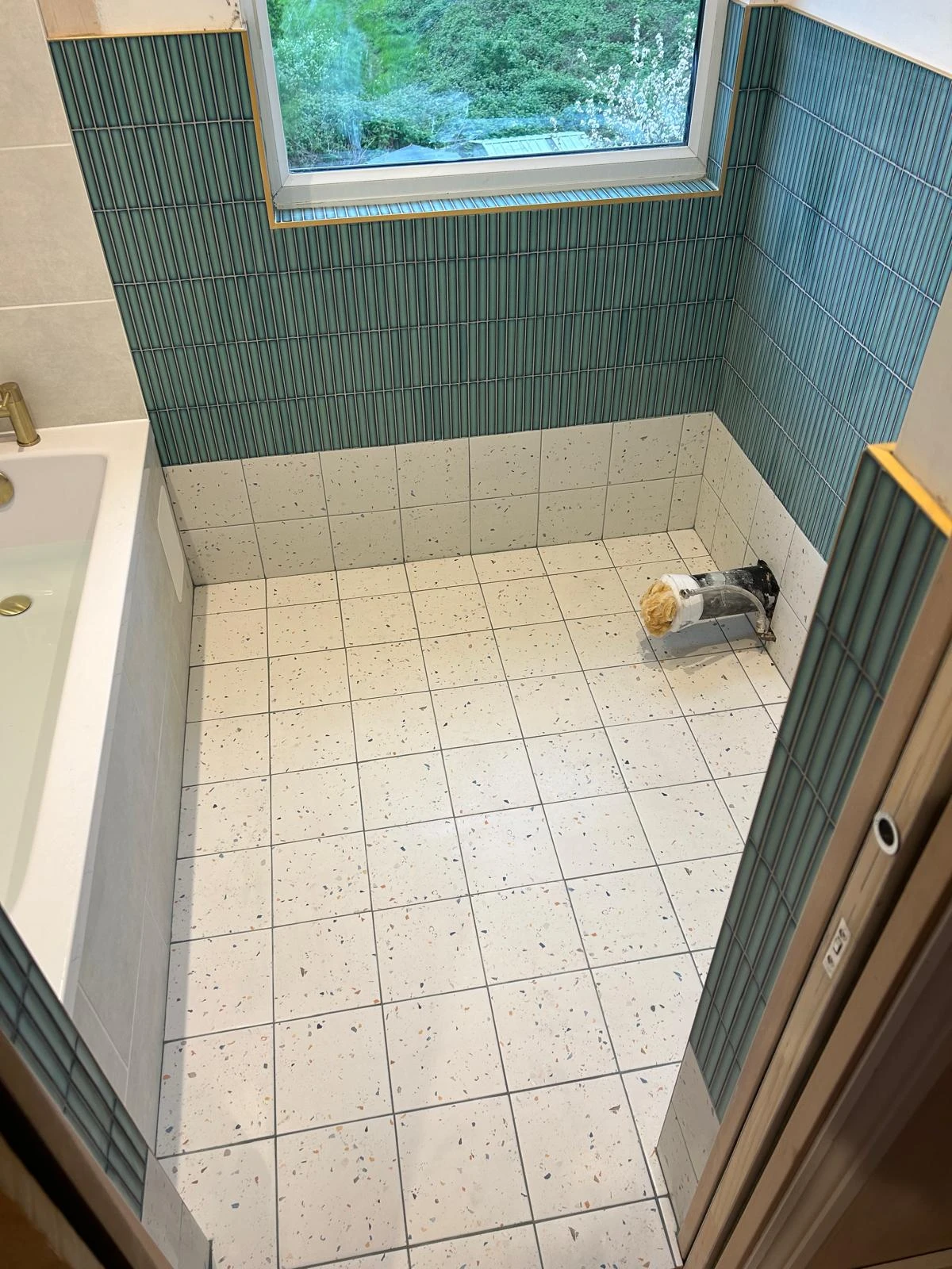 A partially tiled bathroom with a window, featuring green vertical tiles on the walls and speckled white tiles on the floor.