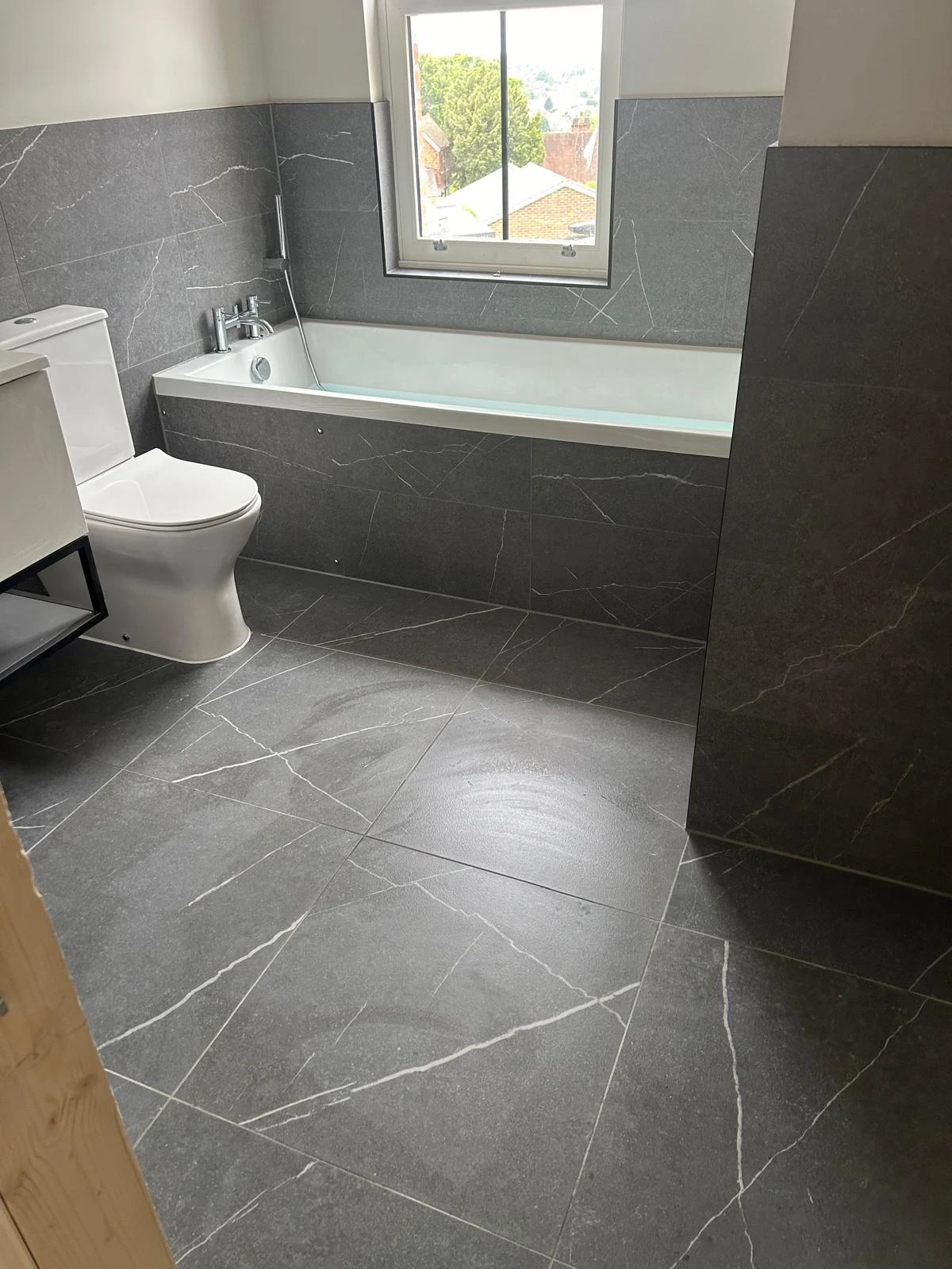 A modern bathroom featuring gray marble tiles, a white toilet, and a bathtub with a window providing natural light.